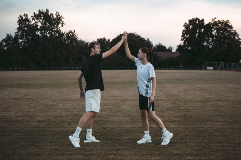 zwei junge sportler in sportkleidung geben sich ein high five auf einem rasenplatz – angst im sport gemeinsam überwinden und mentale stärke aufbauen. das bild transportiert super die botschaft: zusammenhalt, gegenseitige unterstützung und ein positives miteinander im sport, genau der richtige kontrast zum thema angst im sport.