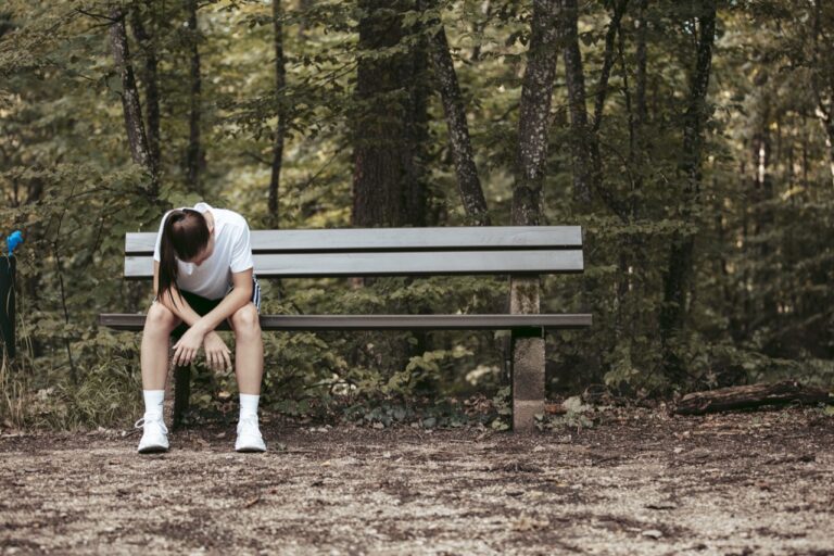 junge sportlerin sitzt erschöpft mit gesenktem kopf auf einer bank im wald, sinnbild für versagensangst im sport